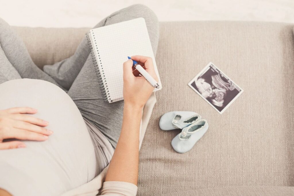 A pregnant person seated on a couch with a note pad and pen for writing her birth plan. A pair of baby booties and ultrasound photo sit next to them.